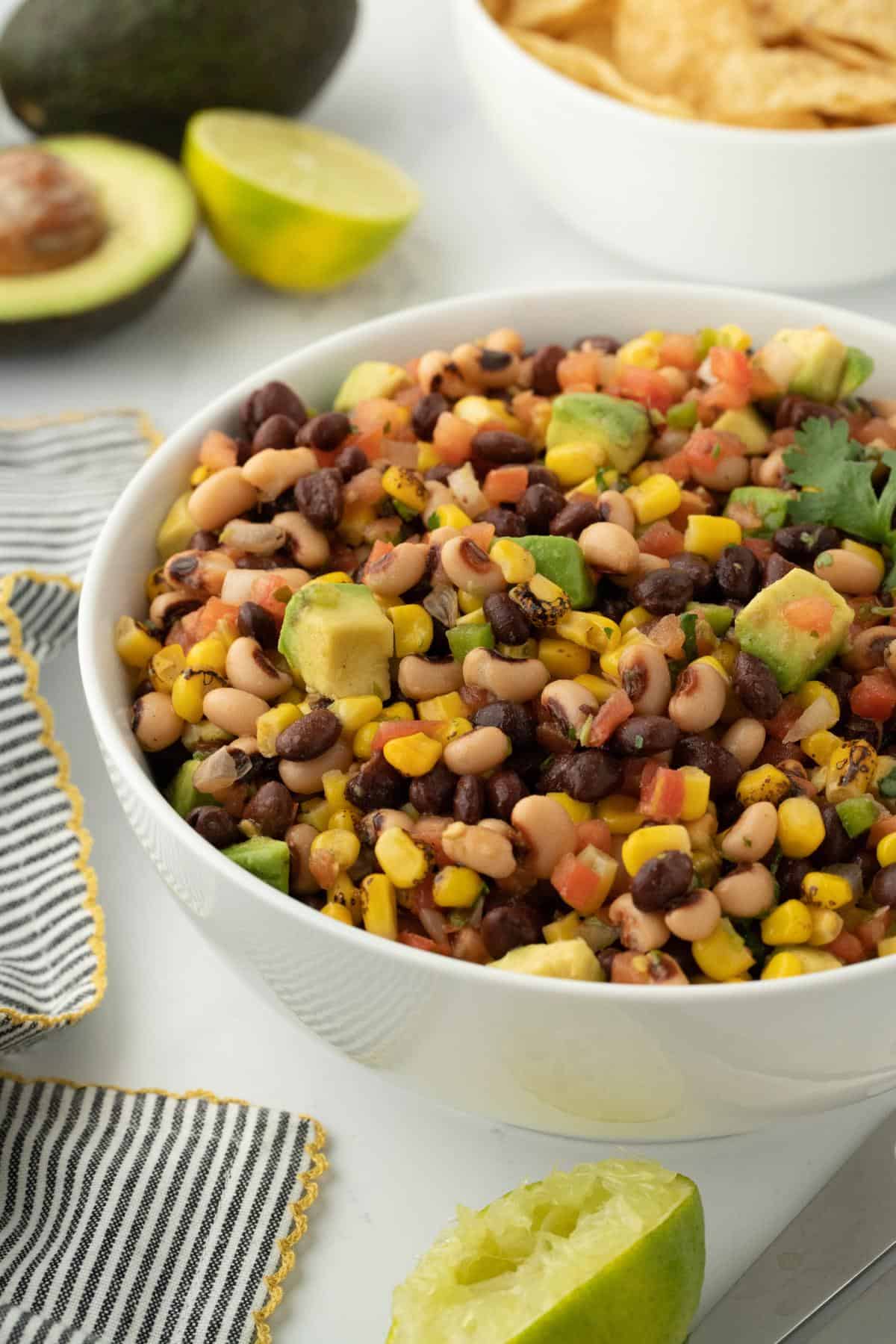 Cowboy caviar in a white bowl with lime wedges, tortilla chips, and avocado slices on the background