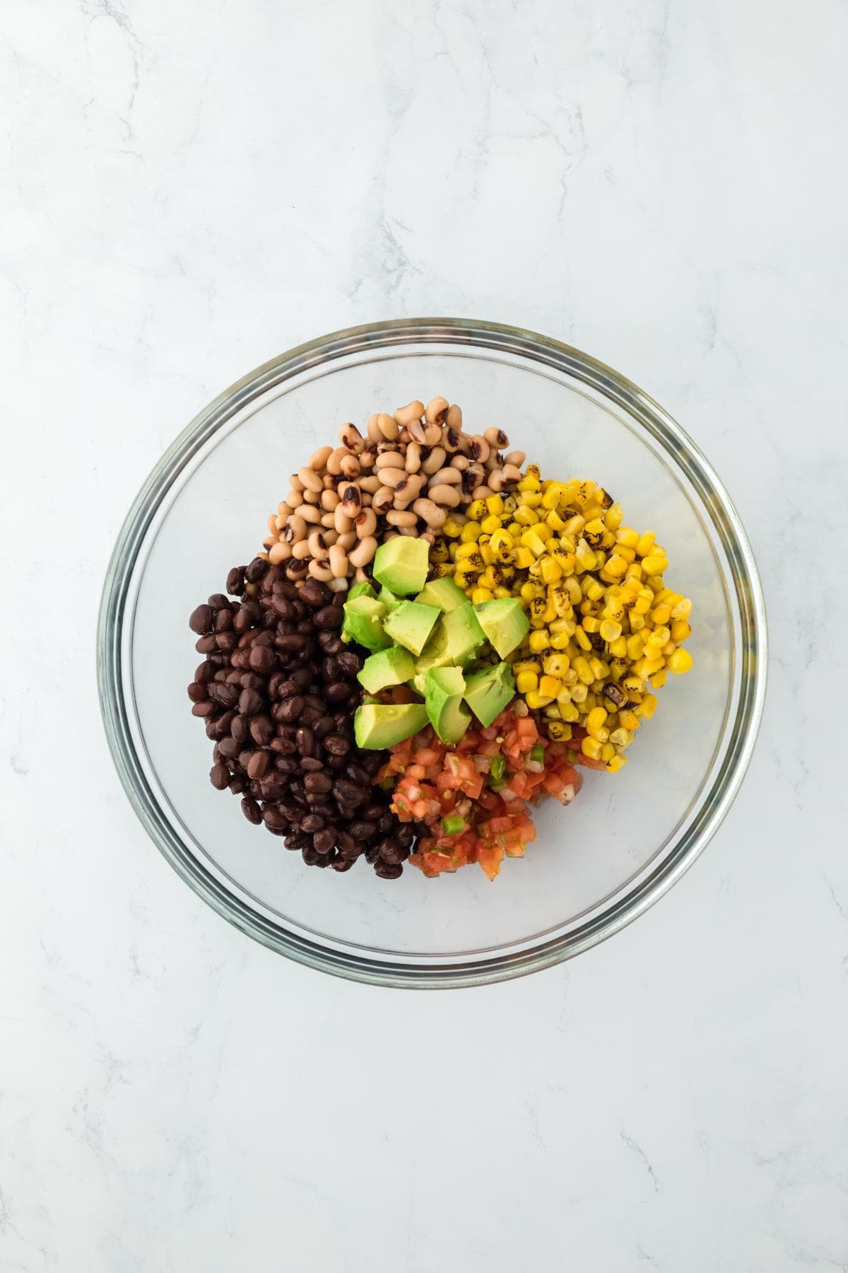 A glass mixing bowl filled with black beans, black-eyed peas, corn, diced avocado, and tomatoes