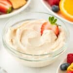 A single bowl of cream cheese fruit dip garnished with a strawberry slice, with fruit and graham crackers in the background