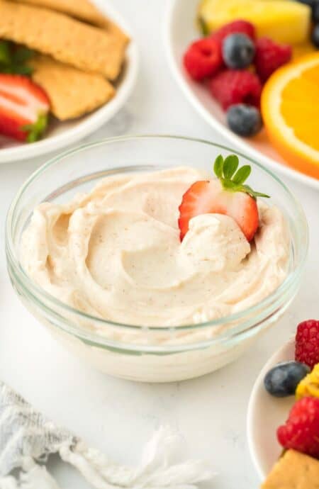 A single bowl of cream cheese fruit dip garnished with a strawberry slice, with fruit and graham crackers in the background