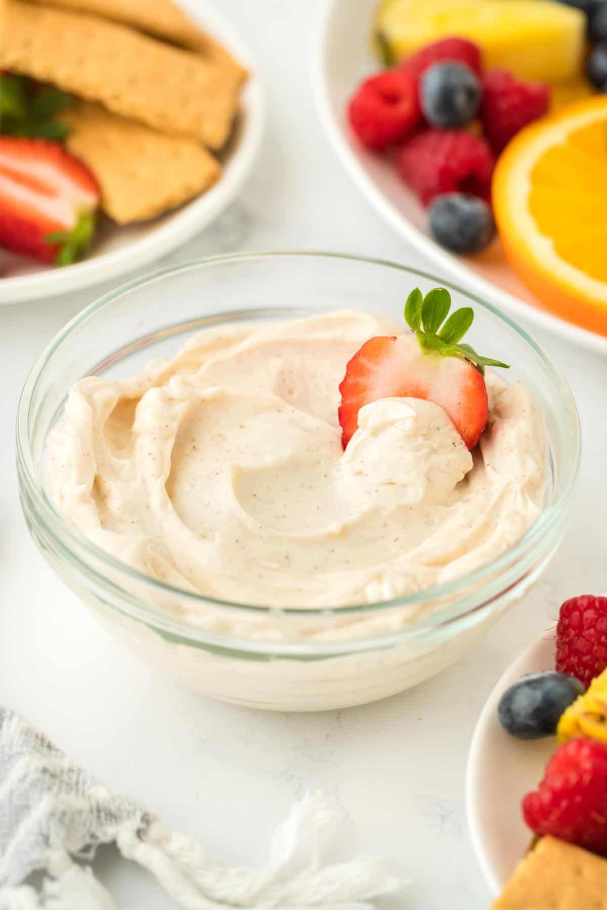 A single bowl of cream cheese fruit dip garnished with a strawberry slice, with fruit and graham crackers in the background