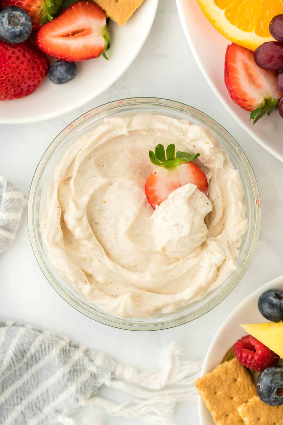 Overhead shot of the cream cheese fruit dip in a glass bowl, topped with a strawberry slice, with fruits like strawberries, blueberries, and pineapple around it