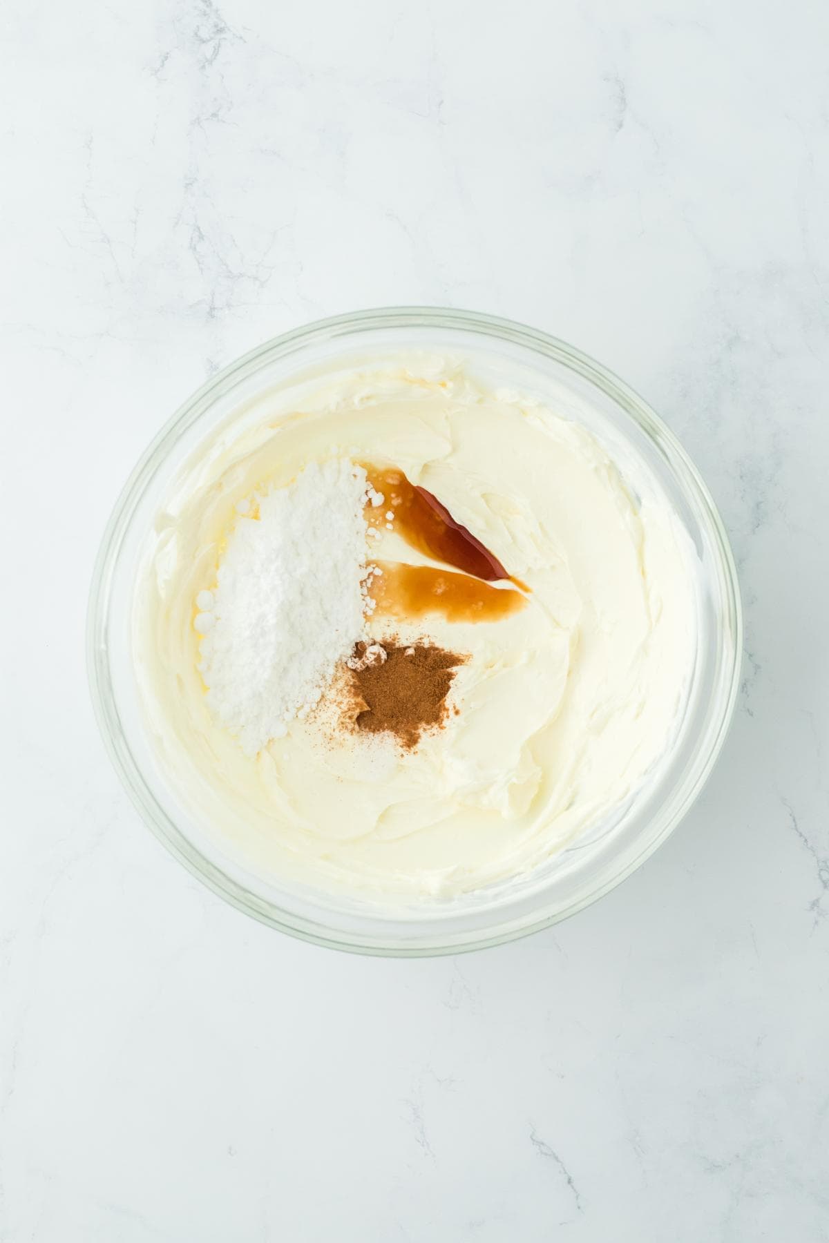 Mixing bowl containing cream cheese, sour cream, powdered sugar, vanilla extract, and cinnamon before being mixed