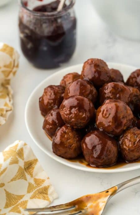 Plate of grape jelly meatballs served with a jar of grape jelly in the background and a fork next to it