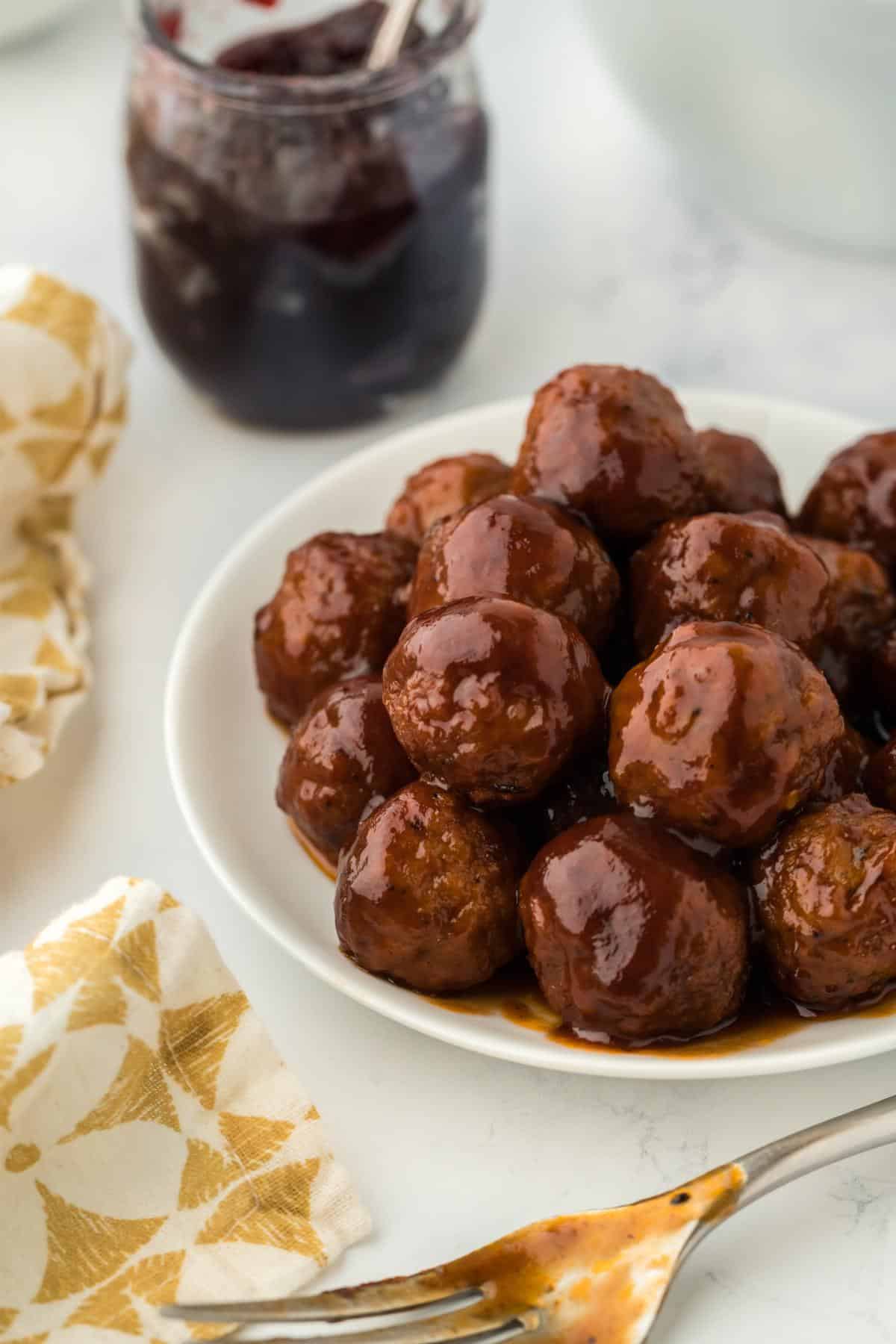 Plate of grape jelly meatballs served with a jar of grape jelly in the background and a fork next to it