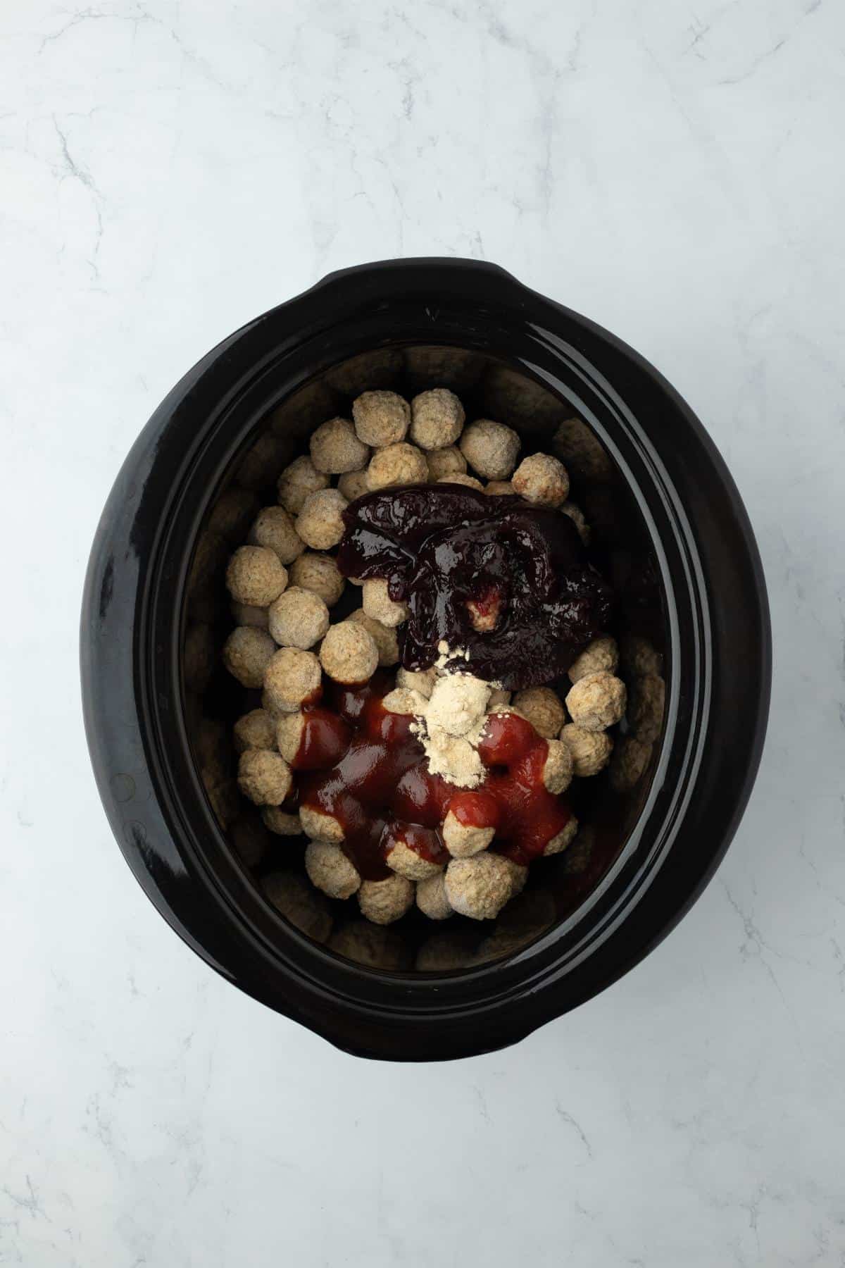 Overhead view of a slow cooker filled with frozen meatballs topped with grape jelly, chili sauce, bbq sauce and garlic powder, ready to be cooked