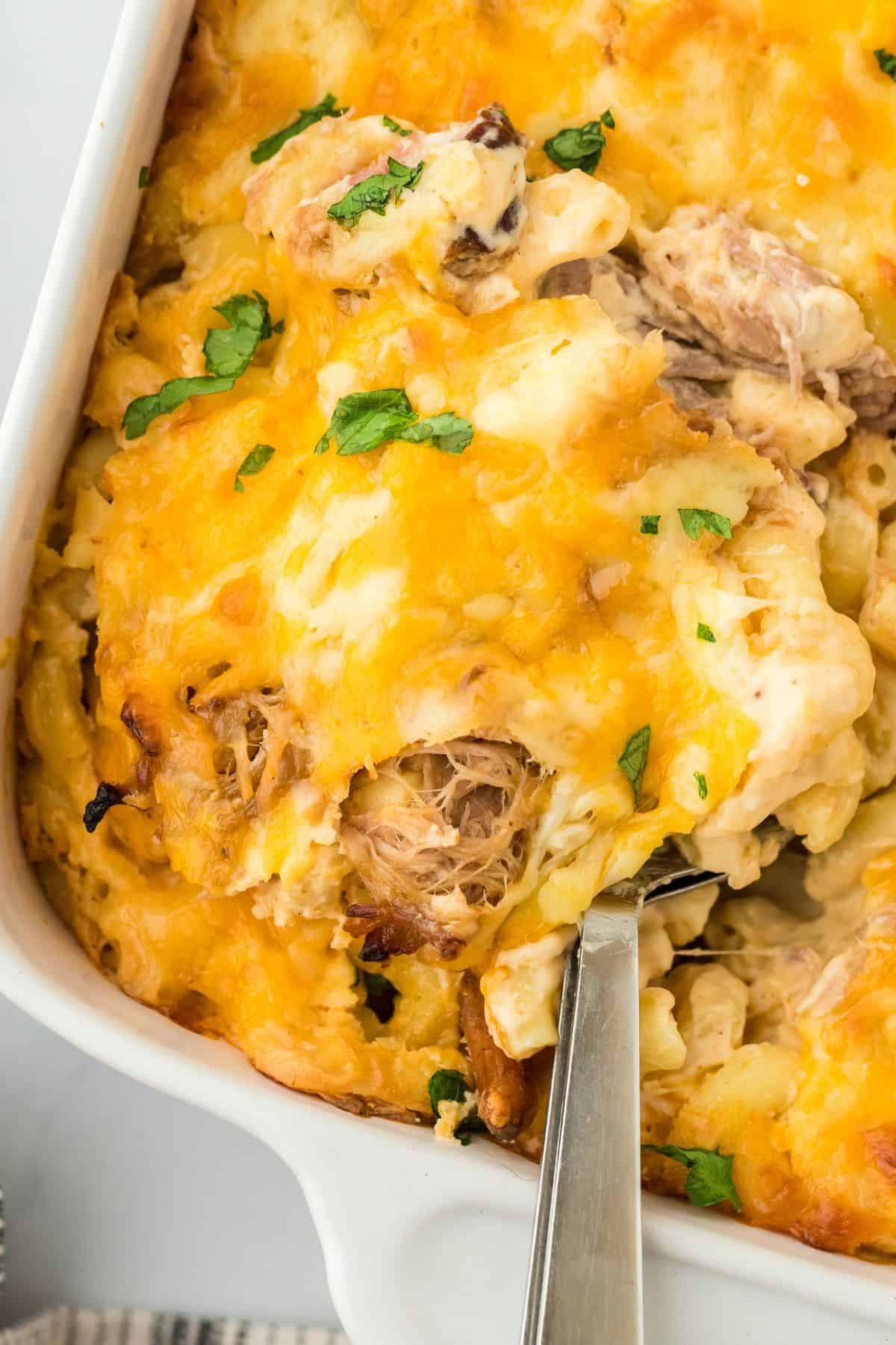 A spoon scooping a serving of the pulled pork mac and cheese off a casserole dish, showing the creamy macaroni and pulled pork filling