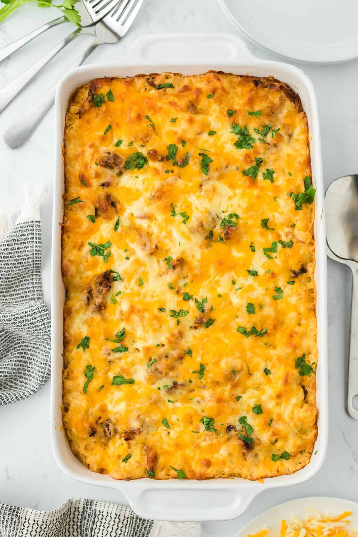 Overhead shot of a casserole dish filled with pulled pork mac and cheese topped with melted cheese and garnished with fresh parsley. The dish is set on a white marble surface with utensils nearby