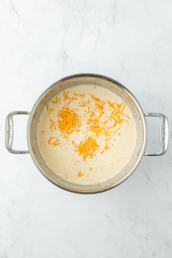 A pot with creamy cheese sauce and shredded cheese being added on top