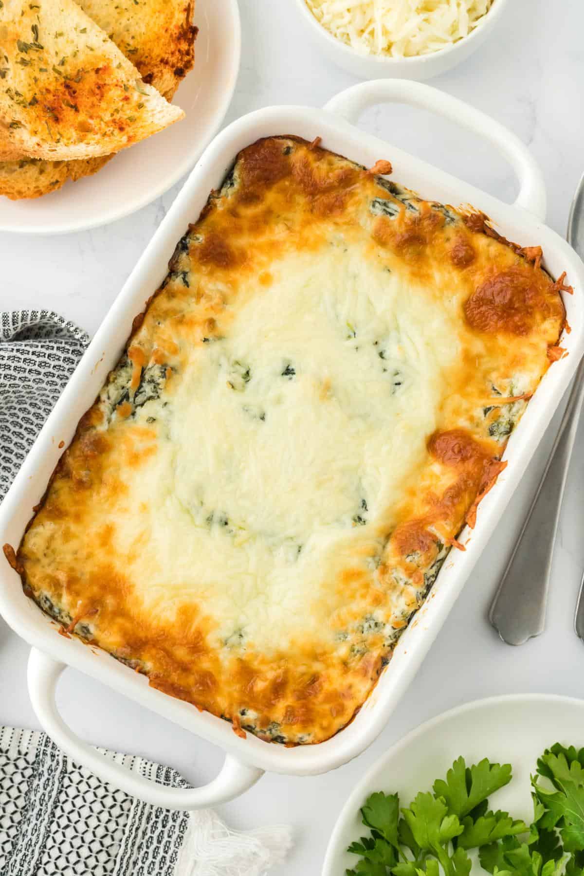 Fully baked spinach dip in a casserole dish with golden, bubbly cheese topping, with more spinach and garlic bread next to it