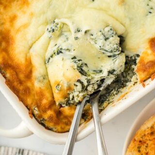 Closeup of a serving of baked spinach dip with two spoons in a white casserole dish, showing melted cheese on top and creamy texture