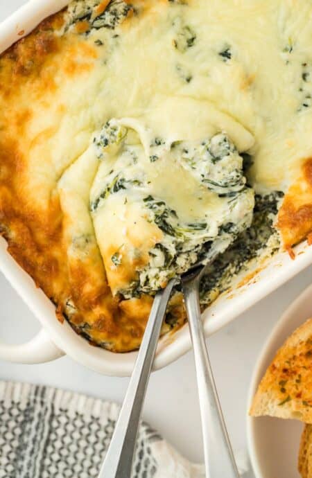 Closeup of a serving of baked spinach dip with two spoons in a white casserole dish, showing melted cheese on top and creamy texture