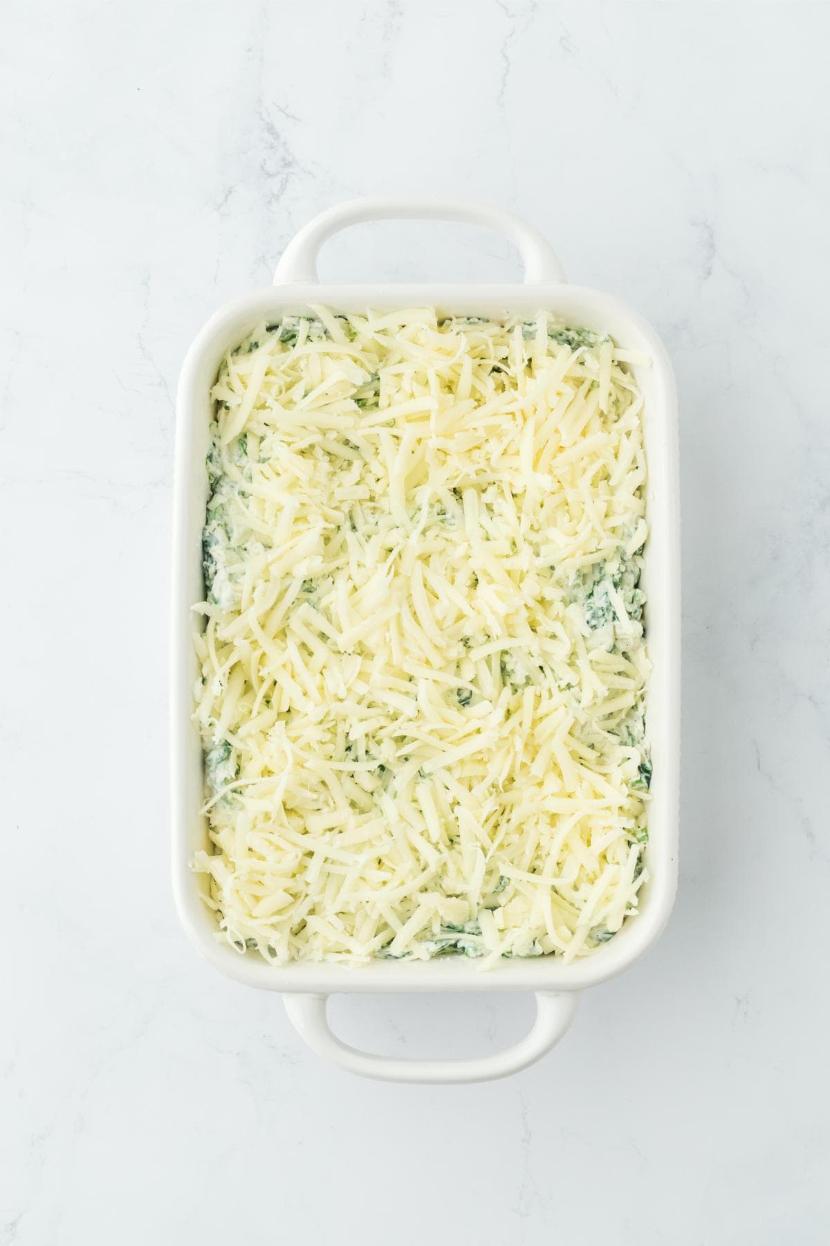 Casserole dish with spinach dip topped with shredded cheese, before baking