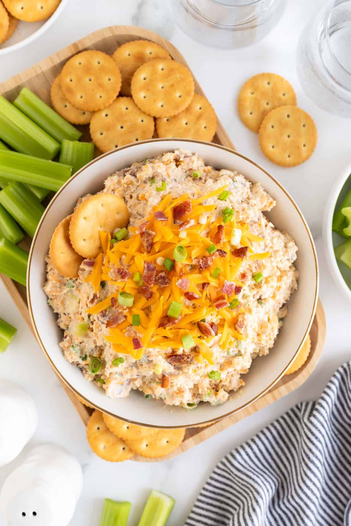 A bowl of creamy million dollar dip topped with crackers, cheddar cheese, bacon bits, and green onions, surrounded by crackers and celery sticks on a wooden tray