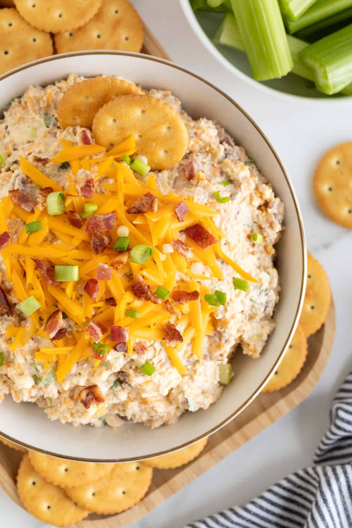 Overhead shot of the million dollar dip highlighting the cheddar cheese, bacon bits, and green onion toppings, with crackers and celery in the background