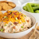 A bowl of creamy million dollar dip topped with cheddar cheese, bacon bits, and green onions, surrounded by crackers and celery sticks on a wooden tray