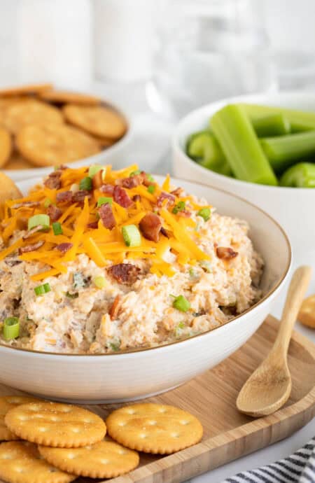 A bowl of creamy million dollar dip topped with cheddar cheese, bacon bits, and green onions, surrounded by crackers and celery sticks on a wooden tray