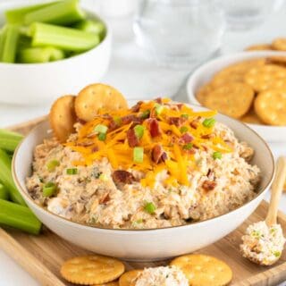 A bowl of creamy million dollar dip topped with crackers, cheddar cheese, bacon bits, and green onions, surrounded by crackers and celery sticks on a wooden tray