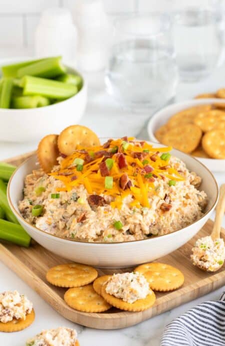 A bowl of creamy million dollar dip topped with crackers, cheddar cheese, bacon bits, and green onions, surrounded by crackers and celery sticks on a wooden tray