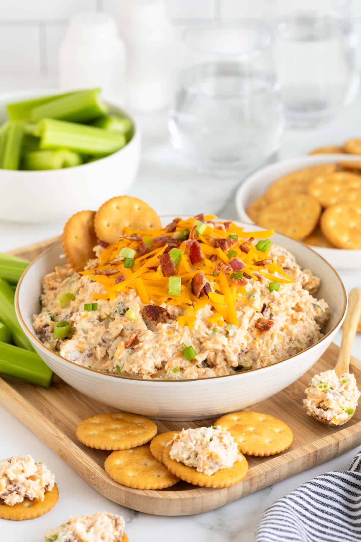 A bowl of creamy million dollar dip topped with crackers, cheddar cheese, bacon bits, and green onions, surrounded by crackers and celery sticks on a wooden tray