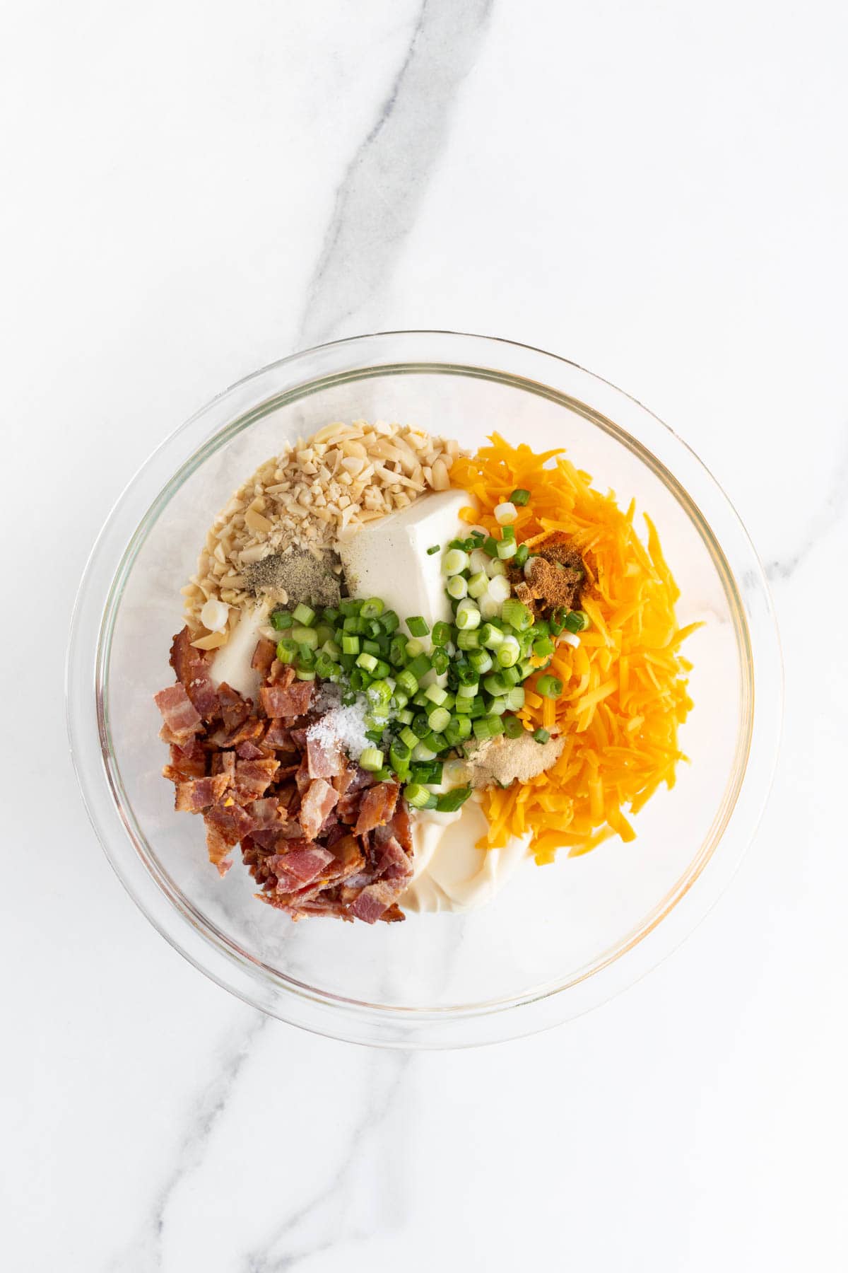 A mixing bowl with dip ingredients such as cheddar cheese, bacon bits, green onions, and cream cheese, arranged before mixing