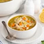 A bowl of chicken and rolled dumplings soup with bright orange carrots, a serving spoon, and a plate, with more soup in the background