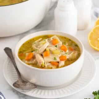 A bowl of chicken and rolled dumplings soup with bright orange carrots, a serving spoon, and a plate, with more soup in the background