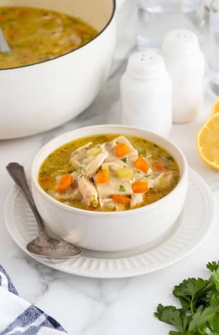 A bowl of chicken and rolled dumplings soup with bright orange carrots, a serving spoon, and a plate, with more soup in the background