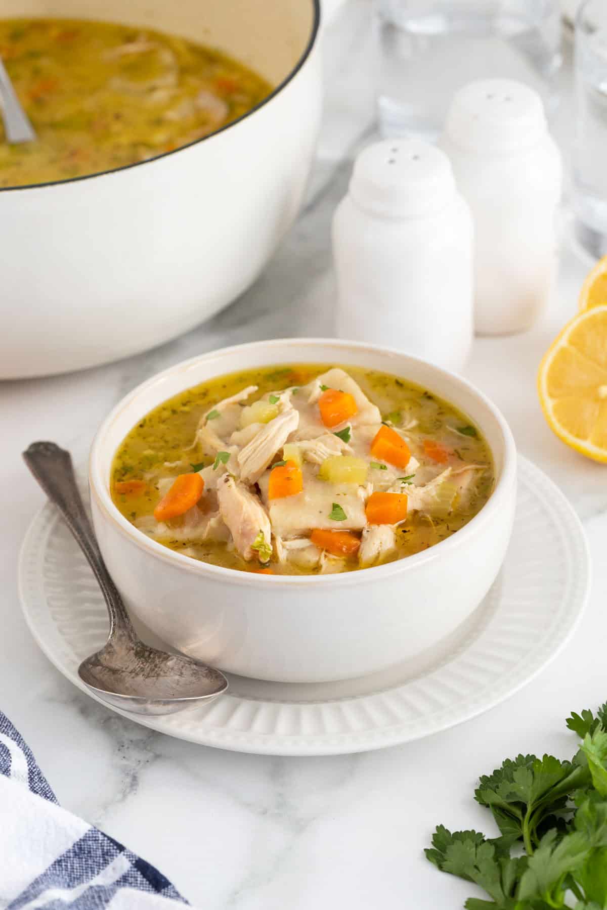 A bowl of chicken and rolled dumplings soup with bright orange carrots, a serving spoon, and a plate, with more soup in the background