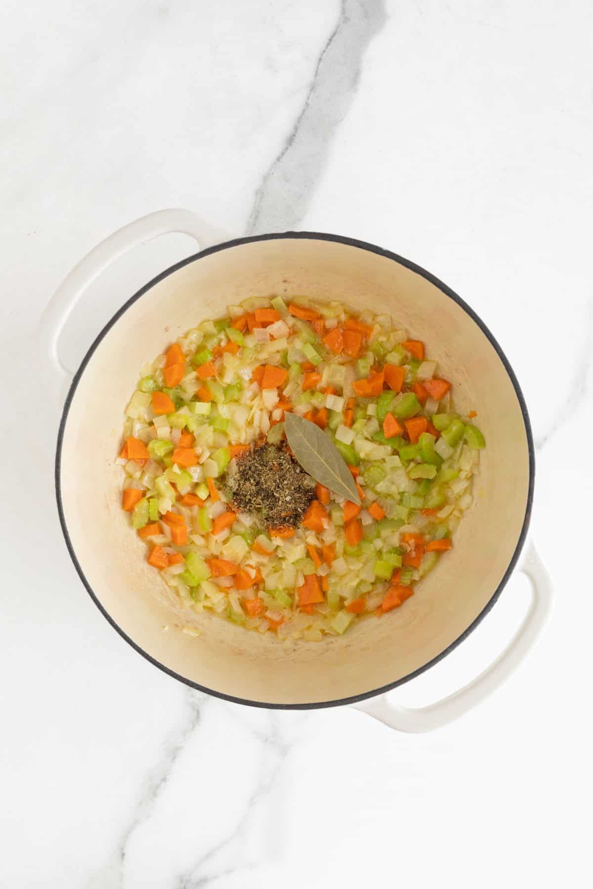 Chopped vegetables (onions, carrots, celery) cooking in a pot with seasonings and a bay leaf