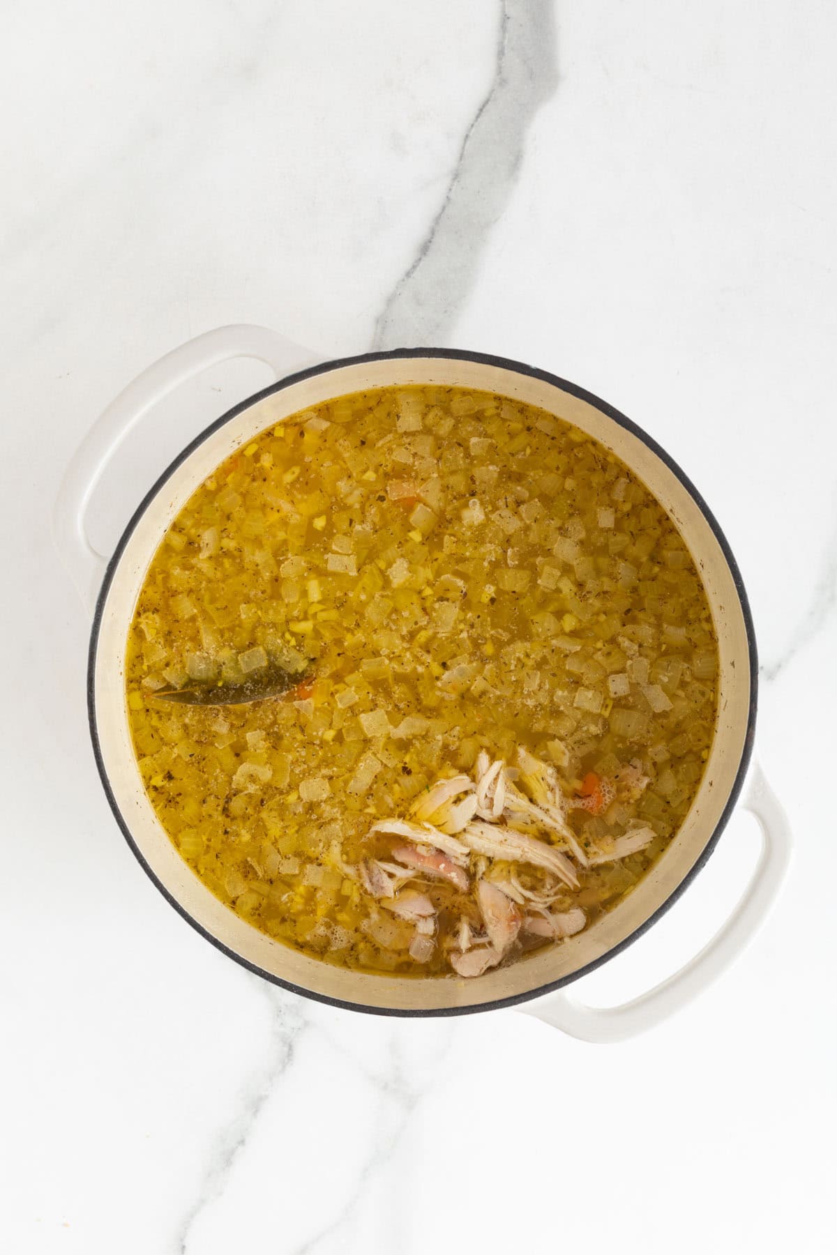 Closeup of a pot filled with a soup or broth with pieces of shredded chicken, vegetables, and seasonings floating inside