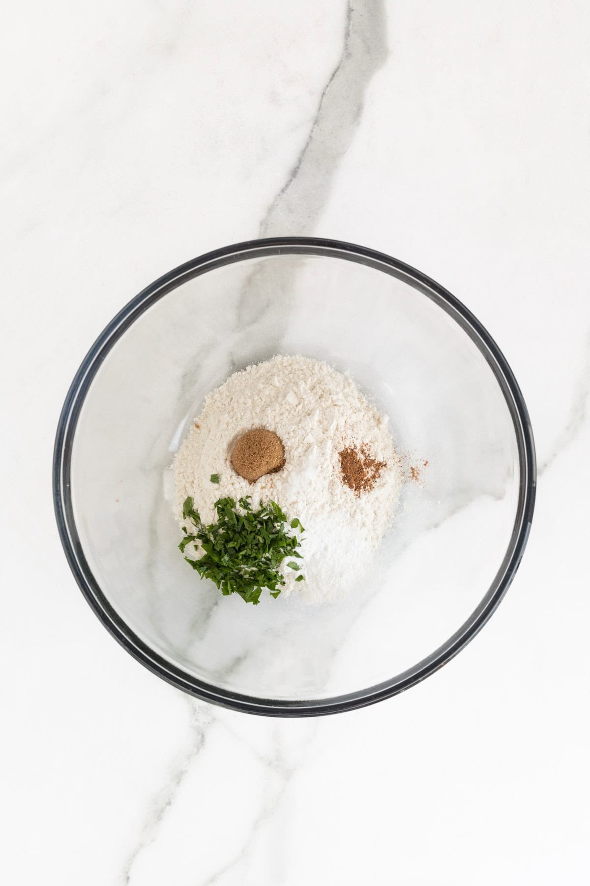 A bowl with flour, brown sugar, spices, and chopped herbs for dumplings, prepared in a mixing bowl