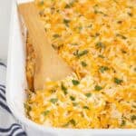 Closeup of a cheesy hashbrown casserole in a white baking dish with a wooden spoon scooping a portion