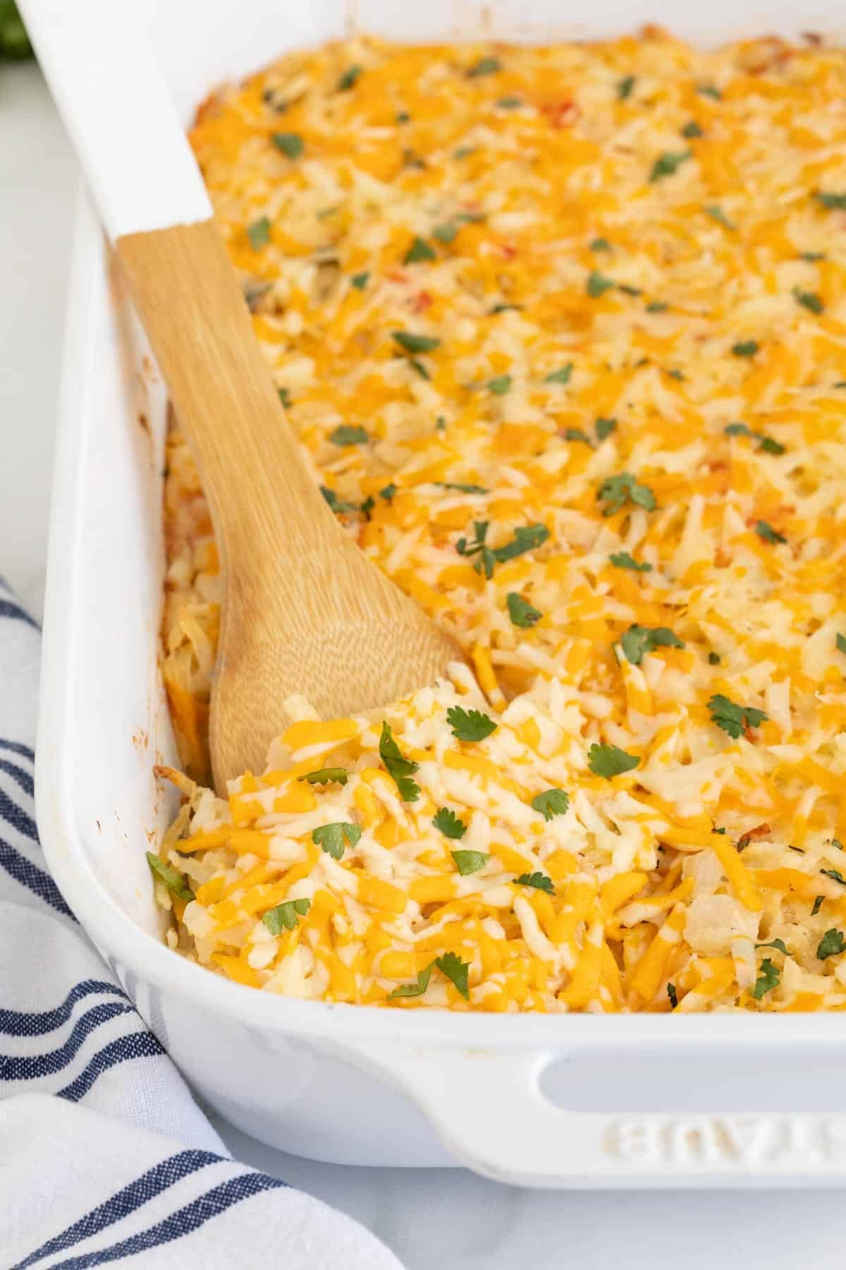 Closeup of a cheesy hashbrown casserole in a white baking dish with a wooden spoon scooping a portion