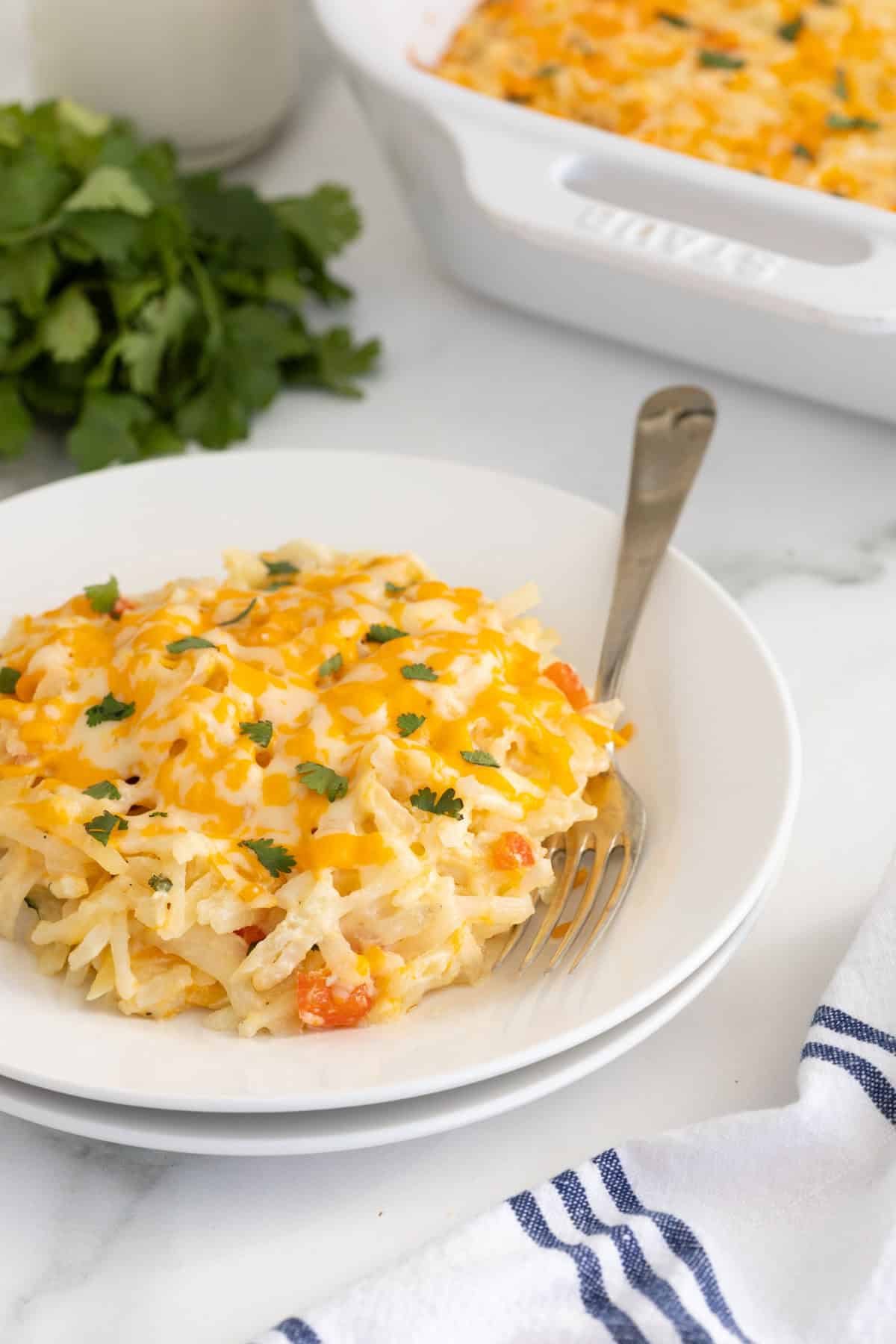 A serving of hashbrown casserole on a white plate with a fork, with a white baking dish in the background with more casserole