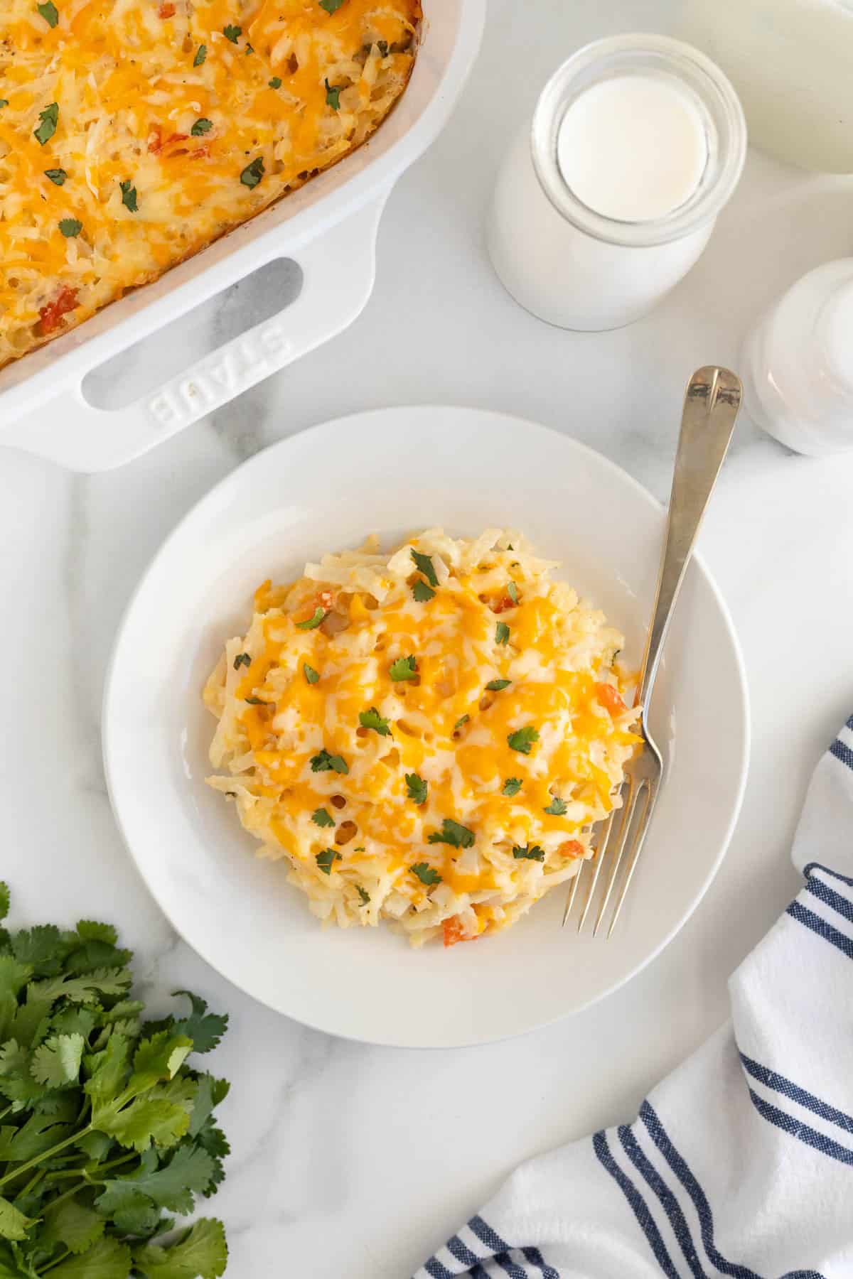 Overhead shot of a plate of hashbrown casserole with a baking dish, a bottle of milk, and fresh parsley surrounding it