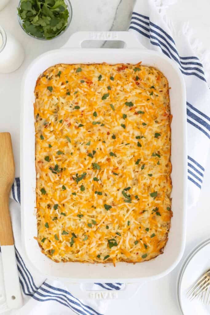 Hashbrown casserole on a marble countertop, surrounded by a blue-striped napkin, fresh herbs, and a milk jar