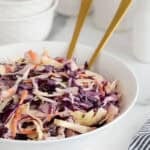 A large white bowl filled with classic coleslaw, with two gold serving spoons sticking out from the bowl