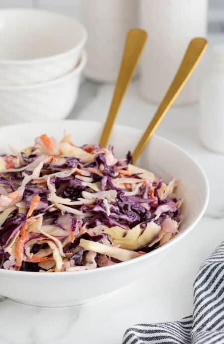 A large white bowl filled with classic coleslaw, with two gold serving spoons sticking out from the bowl