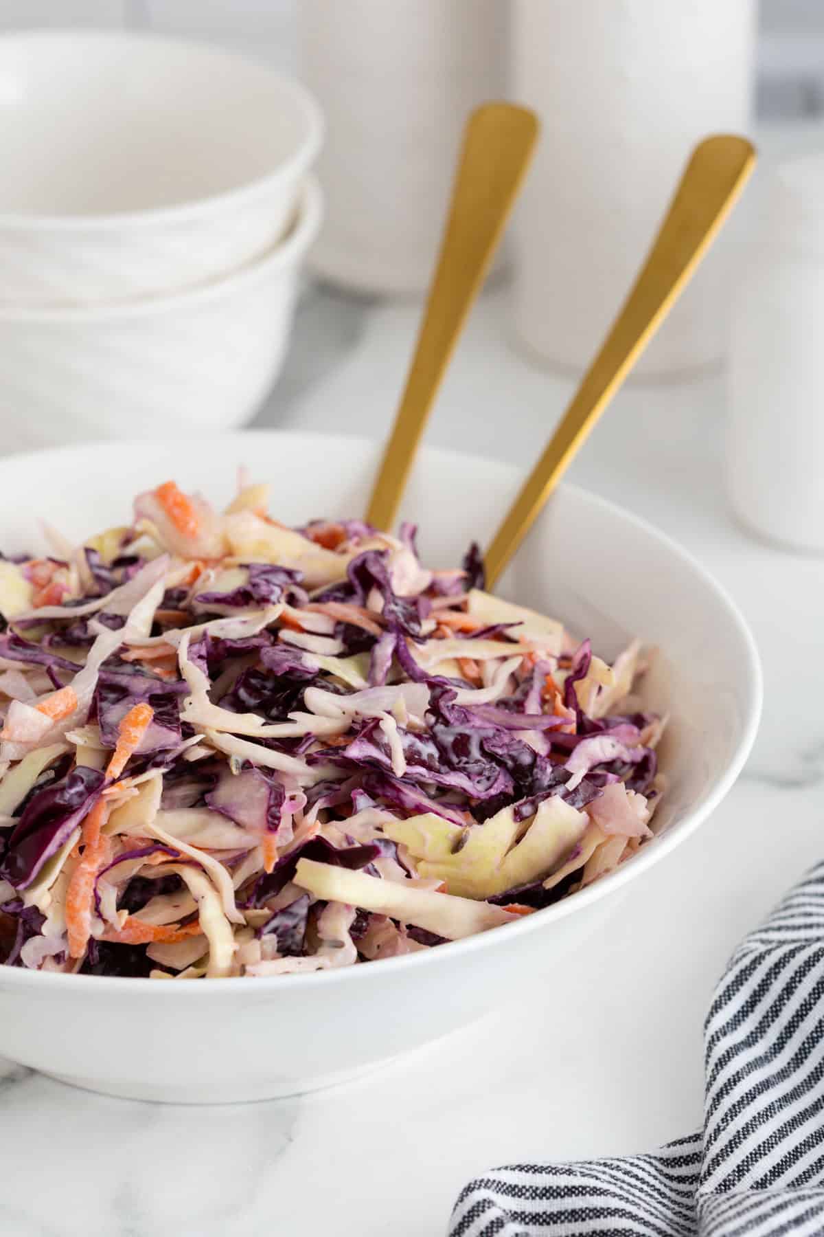 A large white bowl filled with classic coleslaw, with two gold serving spoons sticking out from the bowl