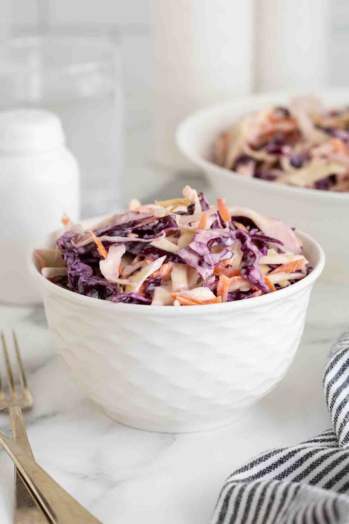 A white textured bowl filled with classic coleslaw, placed on a marble countertop. A larger bowl of coleslaw is visible in the background