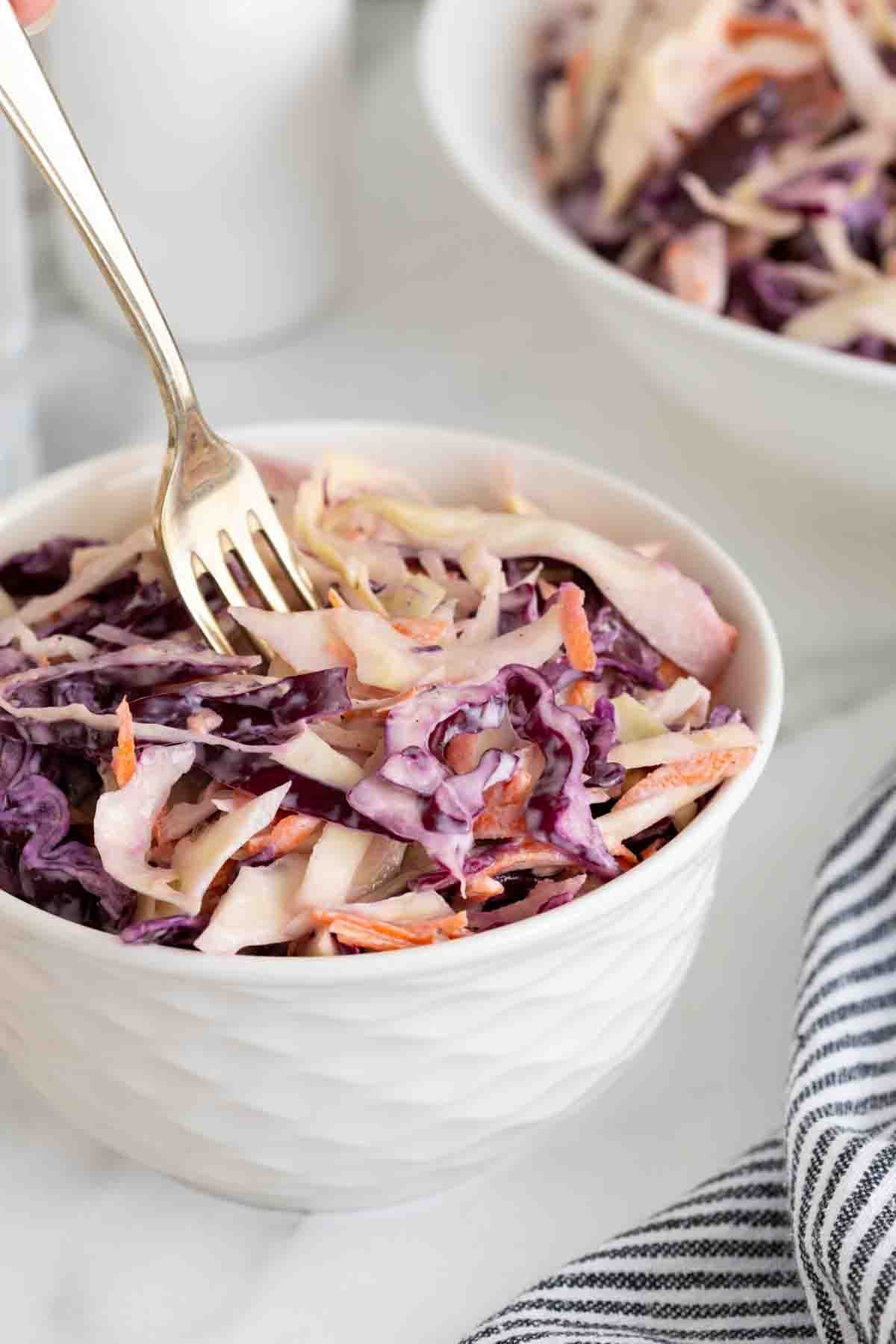 A closeup of a fork resting in a small white bowl of classic coleslaw with more salad in the background