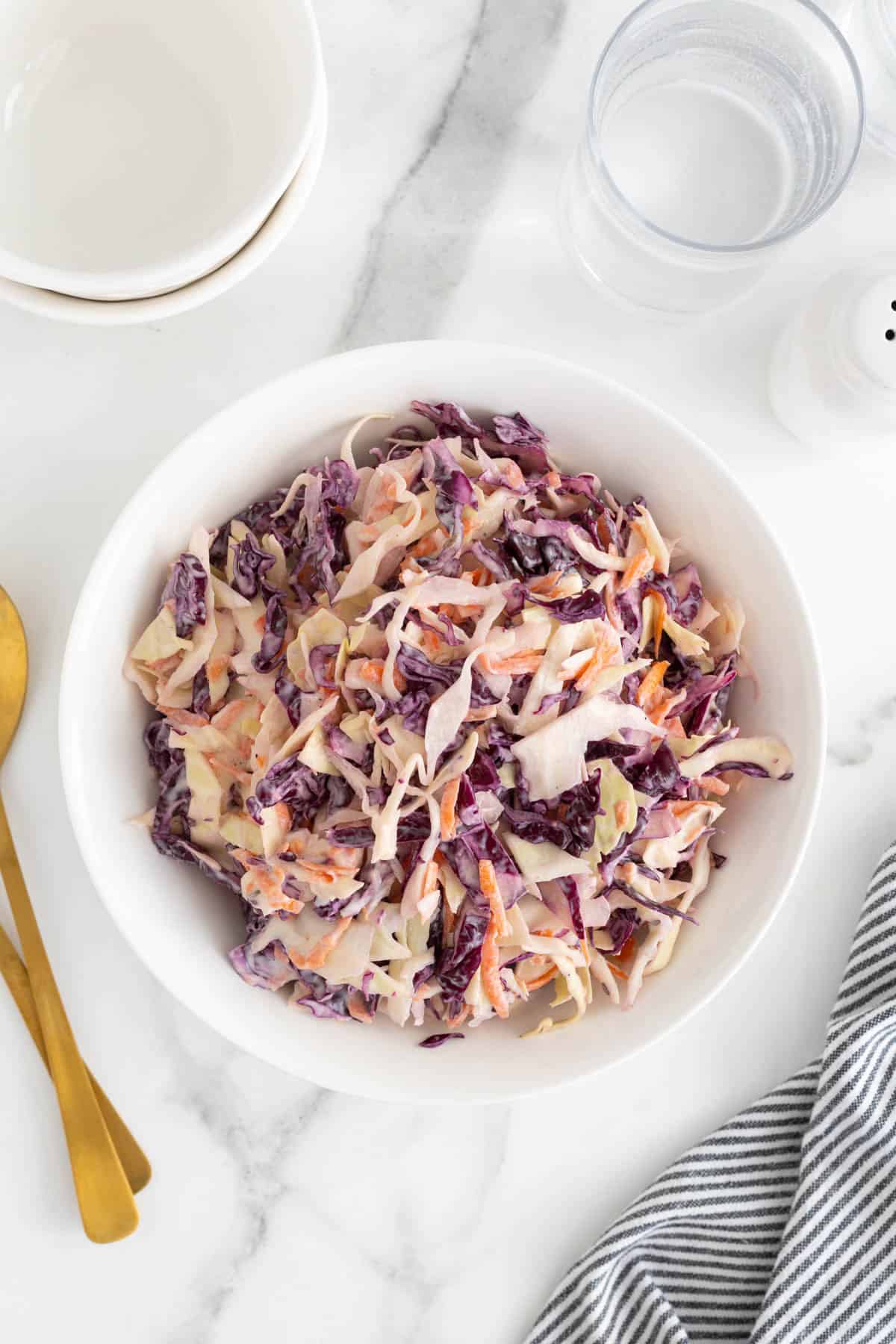 Overhead shot of a large white bowl filled with classic coleslaw, with gold serving spoons, a glass of water and a striped napkin nearby