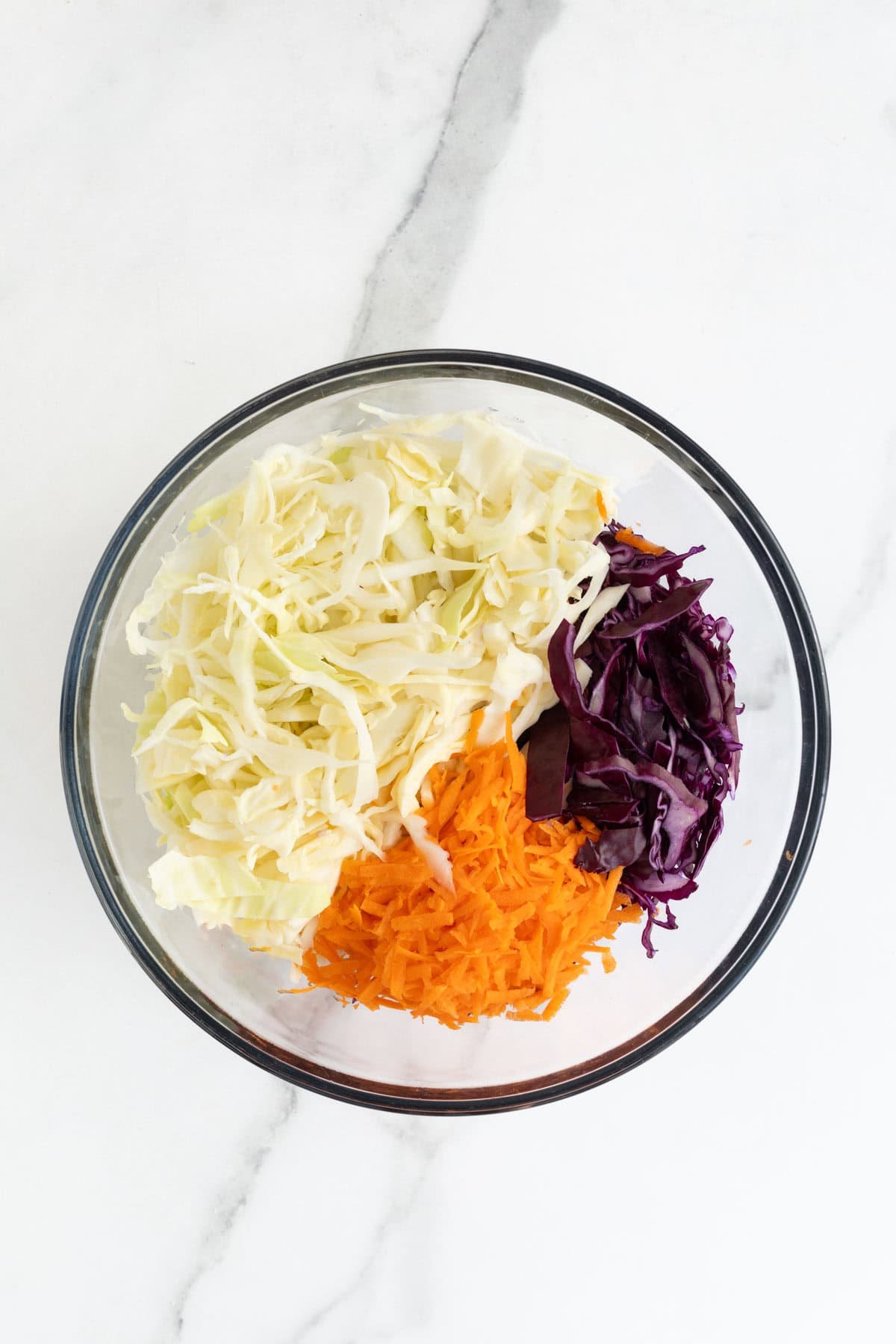 A clear glass mixing bowl with shredded green cabbage, purple cabbage, and grated carrots