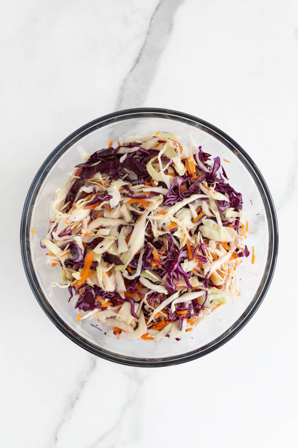 A glass mixing bowl filled with mixed coleslaw ingredients