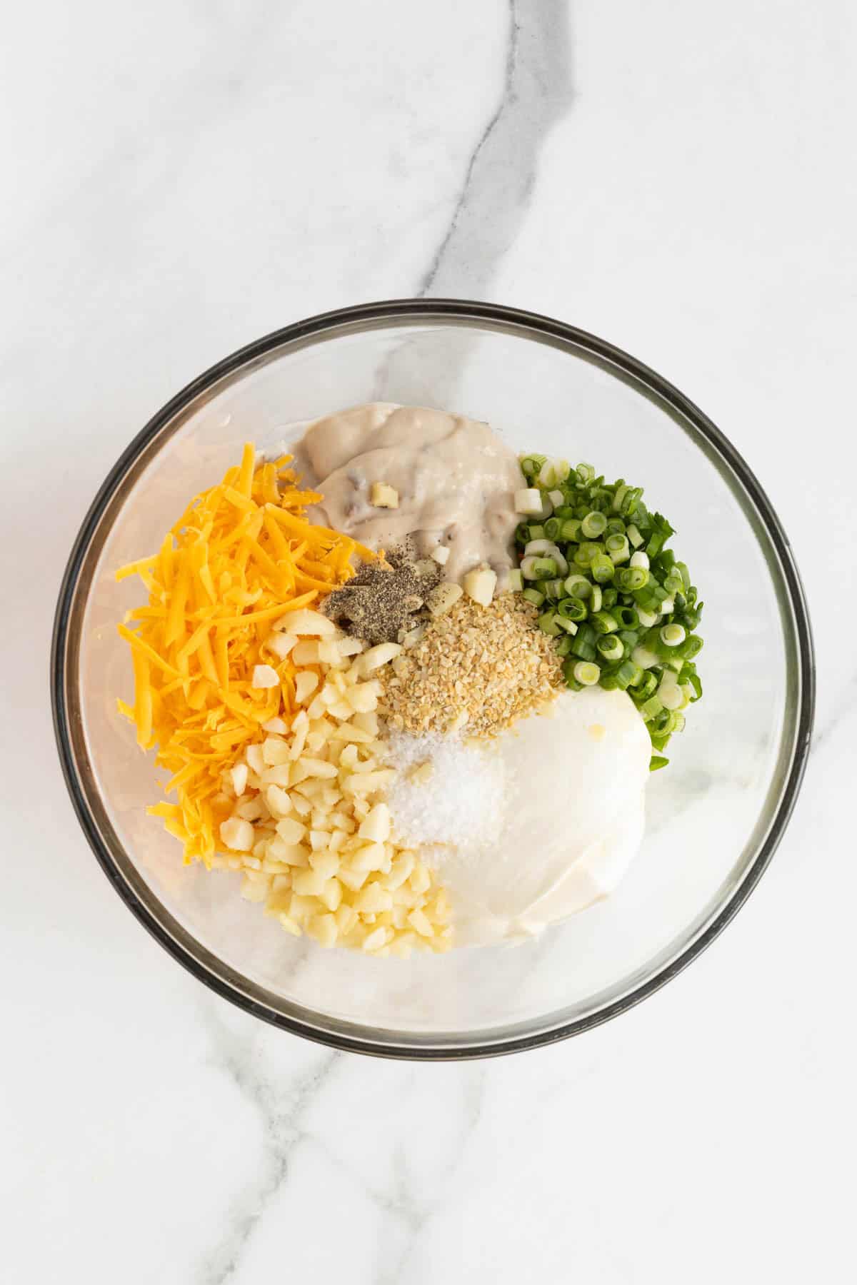 A clear bowl filled with shredded cheddar cheese, sour cream, green onions, cream of mushroom soup, diced potatoes, and seasonings