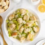 Overhead shot of a Caesar salad in a white bowl with romaine lettuce, shaved Parmesan, and large homemade croutons, surrounded by lemon halves, dressing, and utensils