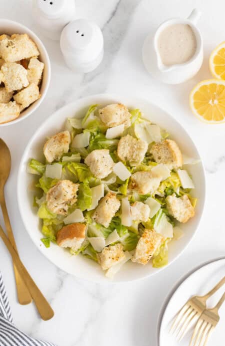 Overhead shot of a Caesar salad in a white bowl with romaine lettuce, shaved Parmesan, and large homemade croutons, surrounded by lemon halves, dressing, and utensils