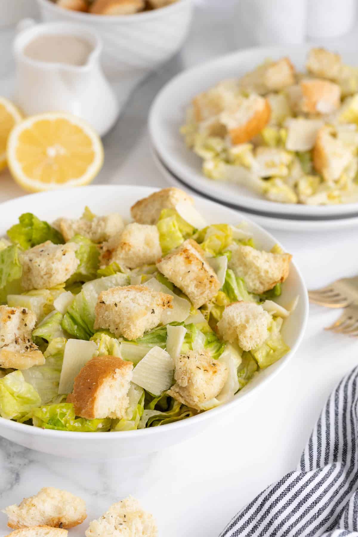Caesar salad on a bowl, with another bowl of salad in the background next to fresh lemon and dressing