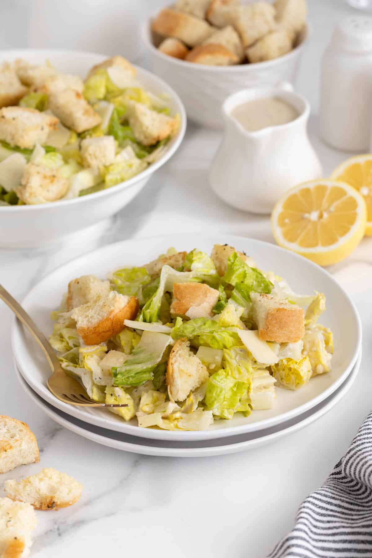 A plated Caesar salad with a gold fork, surrounded by lemons, dressing, another bowl with more salad, and croutons